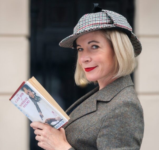 Lucy Worsley in deerstalker hat and book in hand in front of the 221B Baker Street door. (PBS)